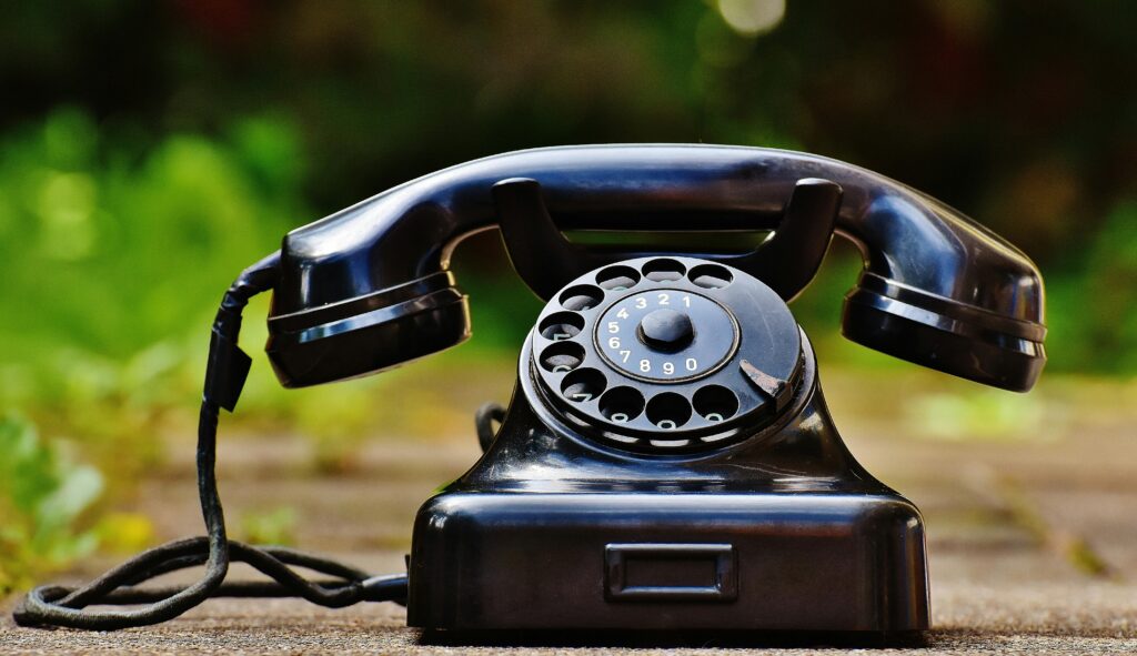 phone-old-year-built-1955-bakelite-163007-163007 Close-up of a classic black rotary phone outdoors with a blurred green background.